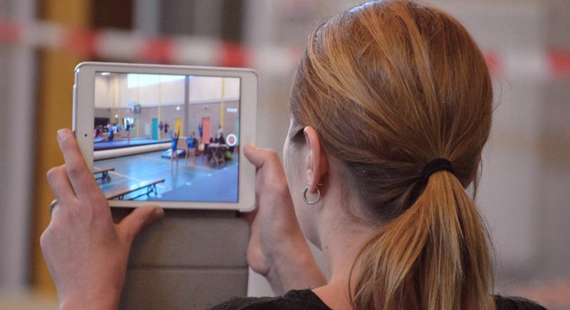 Eine Person mit hellbraunen Haaren, die einen Pferdeschwanz trägt, filmt mit einem Tablet eine Turnveranstaltung in einer Turnhalle. Auf dem Bildschirm sind die turnenden Turner zu sehen, und die Person steht hinter einer Absperrung.