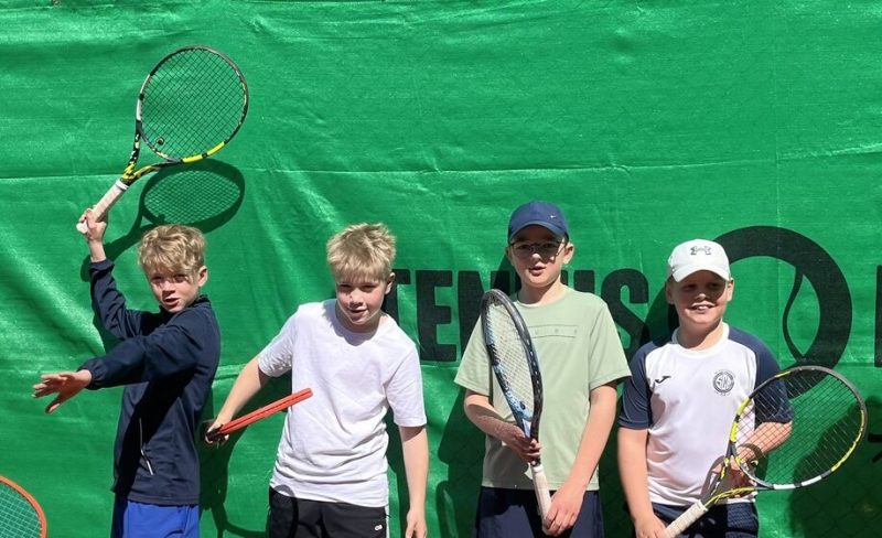 Vier Jungen stehen vor einer grünen Tennisplatzkulisse und halten jeweils einen Tennisschläger in der Hand. Sie tragen Sportkleidung und scheinen an einem sonnigen Tag für ein Foto zu posieren.