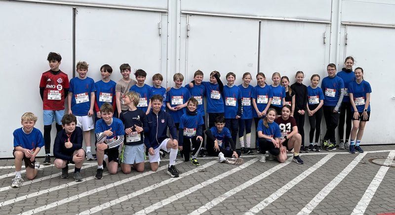 Eine Gruppe von etwa 30 Kindern und Jugendlichen, von denen viele blaue T-Shirts und Startnummern tragen, posiert gemeinsam im Freien vor einer weißen Wand – einige knien, andere stehen – entweder vor oder nach einem Laufwettbewerb.
